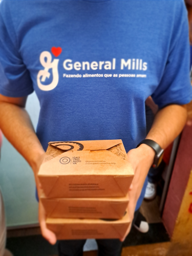 Person holding three stacked brown food boxes, wearing a blue "General Mills" T-shirt.