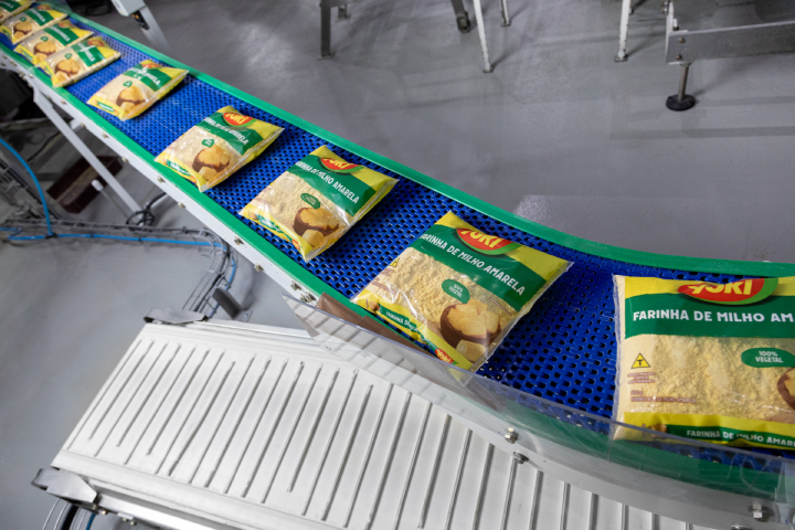 Bags of farinha de milho amarela on a blue conveyor belt in a processing facility.